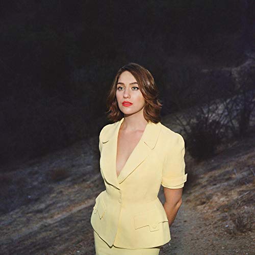 The image depicts a woman in a yellow suit standing on a dirt path. For accessibility purposes, a suitable alt text could be: "Woman in a yellow suit standing outdoors."