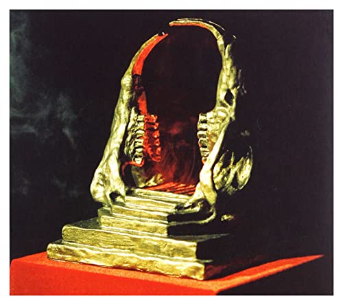 A gold, open-mouthed skull sculpture on a red surface against a black background.
