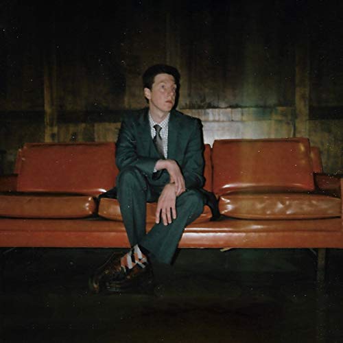 Man in dark suit sits on worn red couch, looking to the right.