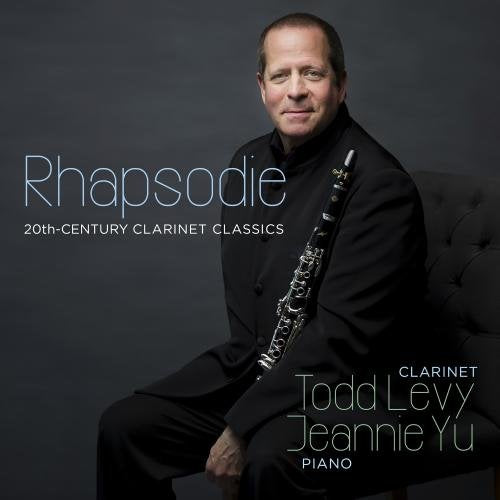 Man in black suit holding clarinet, sitting on a tufted chair, against a dark background.