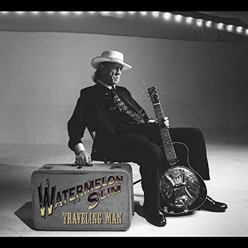 Black and white image of man in suit sitting with guitar and suitcase.