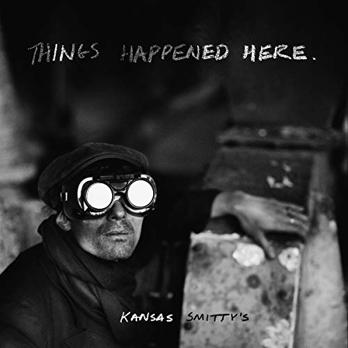 The image is a black-and-white album cover for "Things Happened Here" by Kansas Smitty's. Key Features: * A man wearing goggles and a cap is prominently featured. * The man is positioned on the left side of the image. * The background is dark and blurry, with a metal object visible on the right side. Alt Text: Black-and-white image of a man in goggles and cap, with industrial background.