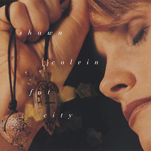 The image depicts a woman holding a pendant necklace, with the text "shawn colvin fat city" overlaid on it. To create an alt text that is concise and descriptive, we can follow these steps: 1. Identify the main subject of the image: The main subject is the woman holding the pendant necklace. 2. Note the key visual elements: The woman's face is partially visible, and she is holding a pendant necklace with a cross-shaped charm. 3. Describe the image in