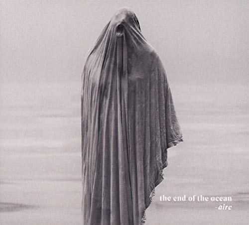 A monochrome image of a person shrouded in fabric, standing on a beach, album cover.