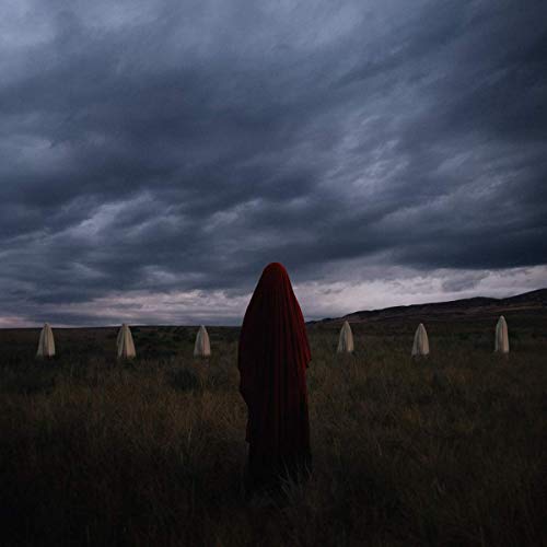 A person in a red hooded cloak stands in a field with white standing stones.