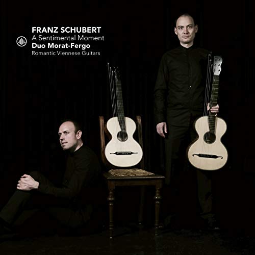 Two men in black, holding acoustic guitars, against a dark background.