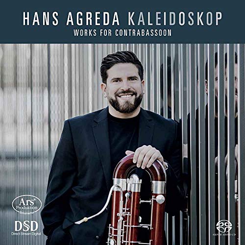A man holding a contrabassoon, standing in front of a metal fence.
