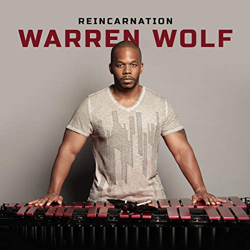 Warren Wolf standing behind vibraphone, wearing a light-colored t-shirt, against a grey background.