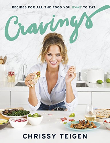 "Chrissy Teigen in a kitchen, holding a taco, surrounded by food on a counter."