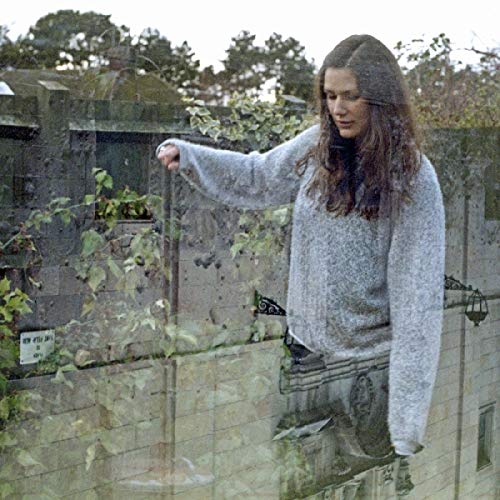 A woman with long brown hair stands against a stone wall, wearing a gray sweater.