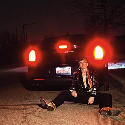 "Man in sequined jacket sits by a car with 'Recovery' license plate at night."