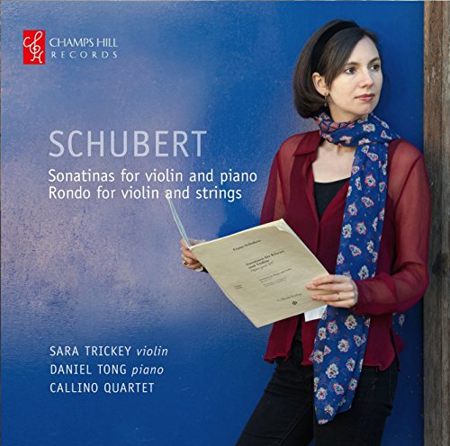 A woman holding sheet music stands against a blue wall, wearing a red cardigan.