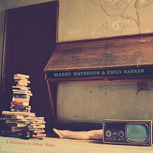 The image depicts a wooden desk with a hinged top, accompanied by a stack of books to its left. The desk features the names "MARRY WATERSON & EMILY B." in white text on the front. For accessibility purposes, a suitable alt text could be: "A wooden desk with a stack of books."
