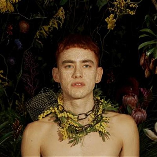 Man with painted face and floral garland around neck, surrounded by foliage.