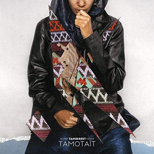 A person in a black leather jacket and patterned scarf stands against a gray wall.