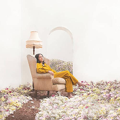 Woman in yellow lounging in beige armchair surrounded by flowers against a white background.