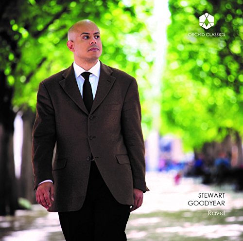 Bald man in brown suit stands outdoors with trees and water in the background.