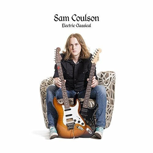 Man with long hair sits holding two electric guitars in a patterned chair.