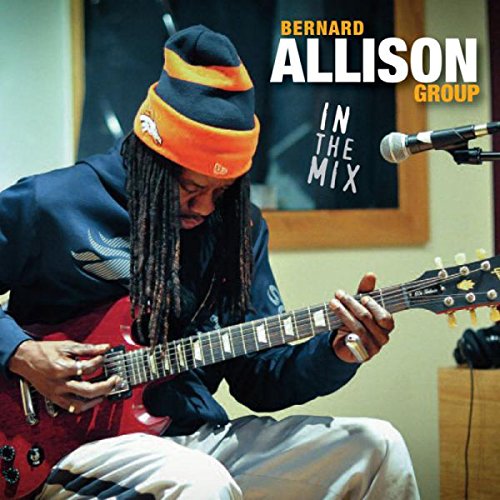 Man playing red guitar, wearing beanie, in a recording studio with microphone nearby.