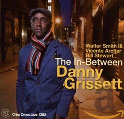 Man in blue suit and striped scarf stands on a dimly lit city street.