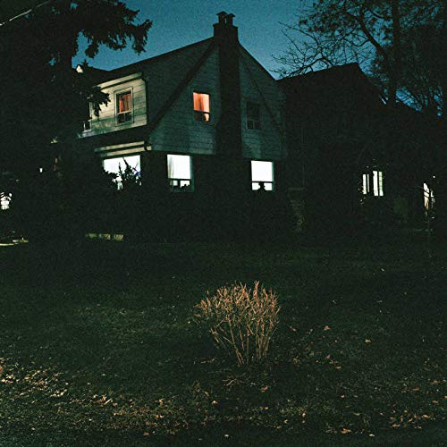 A dark house with lit windows at dusk, surrounded by trees and a yard.