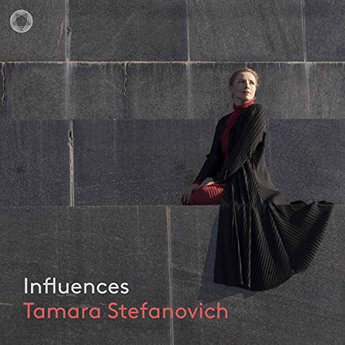 Woman in long black dress holding red clutch, standing against a grey stone wall.