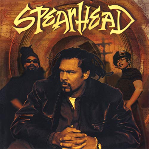 The image depicts a CD cover for the album "Spearhead" by Various, featuring a man in the foreground and two individuals behind him. The man is dressed in a black leather jacket over a white shirt, with his hands clasped together in front of him. He has dark hair and is looking to the left. Behind him, on the left, stands a bearded man wearing a black hat and shirt, while on the right is a woman with short hair, also dressed in black