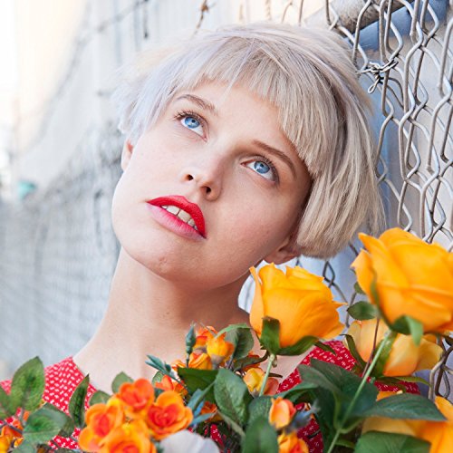 A woman with short blonde hair and red lipstick holds orange roses against a chain-link fence.