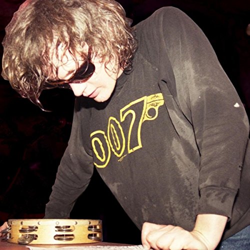 Man in dark shirt with "007" logo plays tambourine on a table.