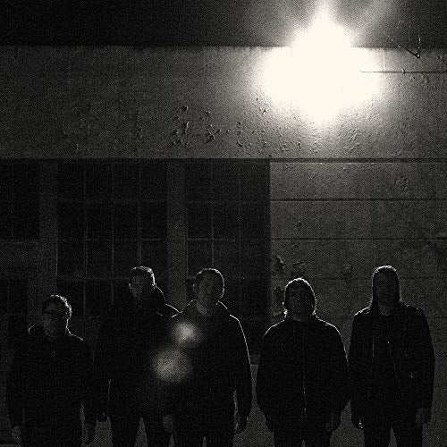 Silhouetted band members stand before a brick wall under a bright streetlight.