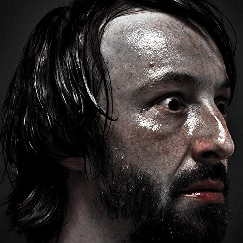 A man with dark hair and a beard, his face painted silver, against a dark background.