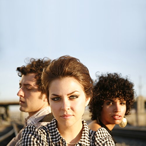 "Three band members, a woman in front and two men behind her, outdoors."