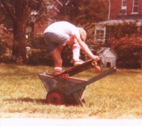 A boy skateboards on a wheelbarrow in a grassy yard with houses in the background.