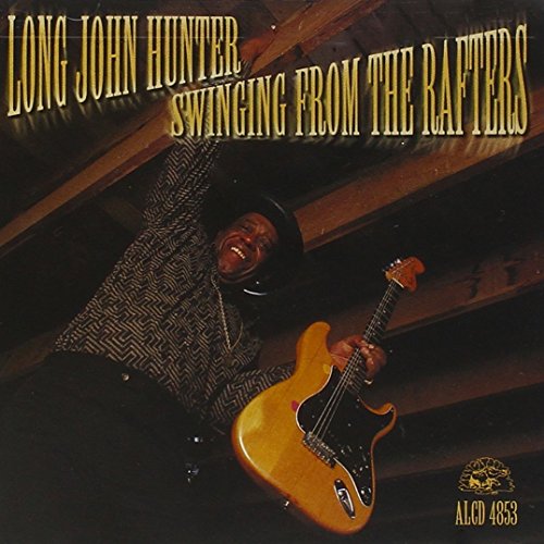 "Album cover: man hanging upside down from rafters, holding a guitar."