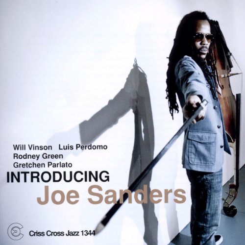 Man with dreadlocks holding a double bass, standing in front of a white wall.