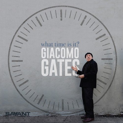 Man in black suit stands beside large clock face on gray wall, holding watch.