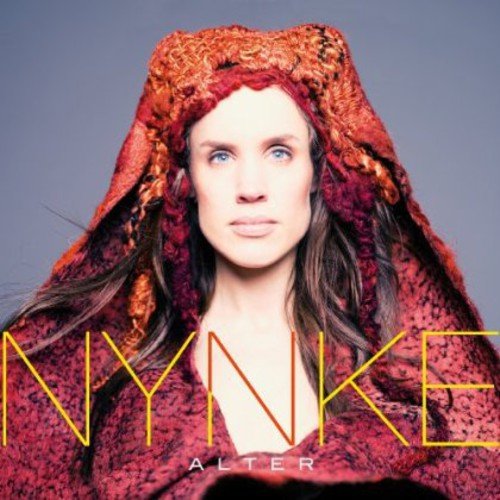 Woman with long brown hair and red patterned hooded cloak, against a gray background.