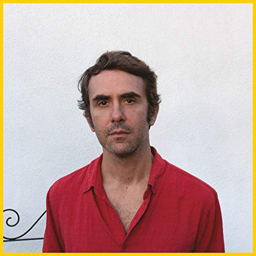 Man with short brown hair and red shirt stands against a white wall.