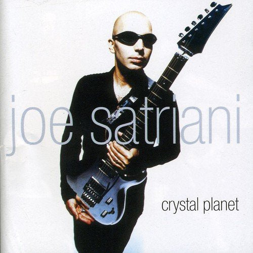 Joe Satriani holding a silver guitar, wearing black, against a white background.
