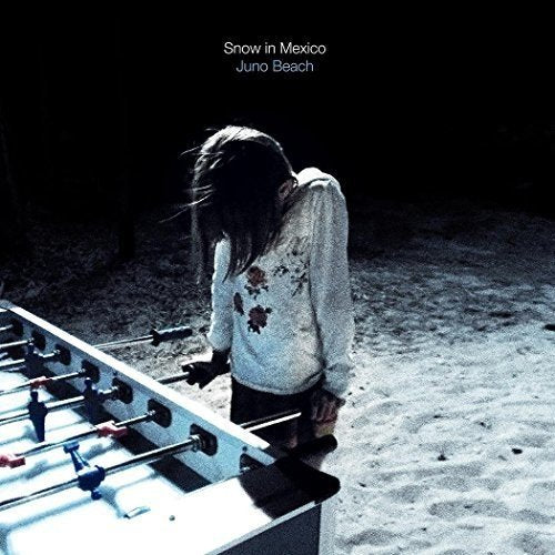 Album cover: a girl standing by a foosball table on a sandy surface.