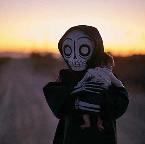A person in a black hooded cloak and skeleton mask holds a baby at sunset.