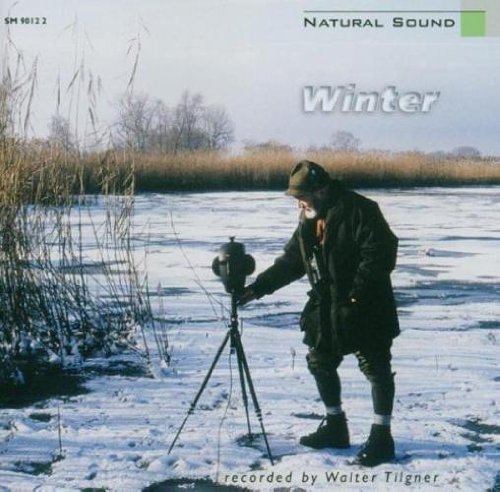 Man in winter clothing stands on frozen lake, adjusting sound recording equipment on tripod.