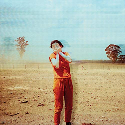A person in orange stands in a dry field, hands covering their face.