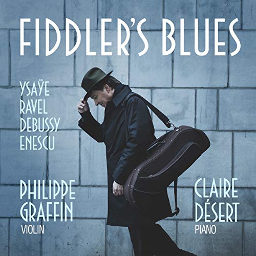 The alt text for the image is: "Man in black coat and hat holding violin case against stone wall."