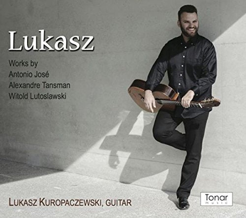 Man in black shirt and pants holding a guitar, standing against a gray wall.