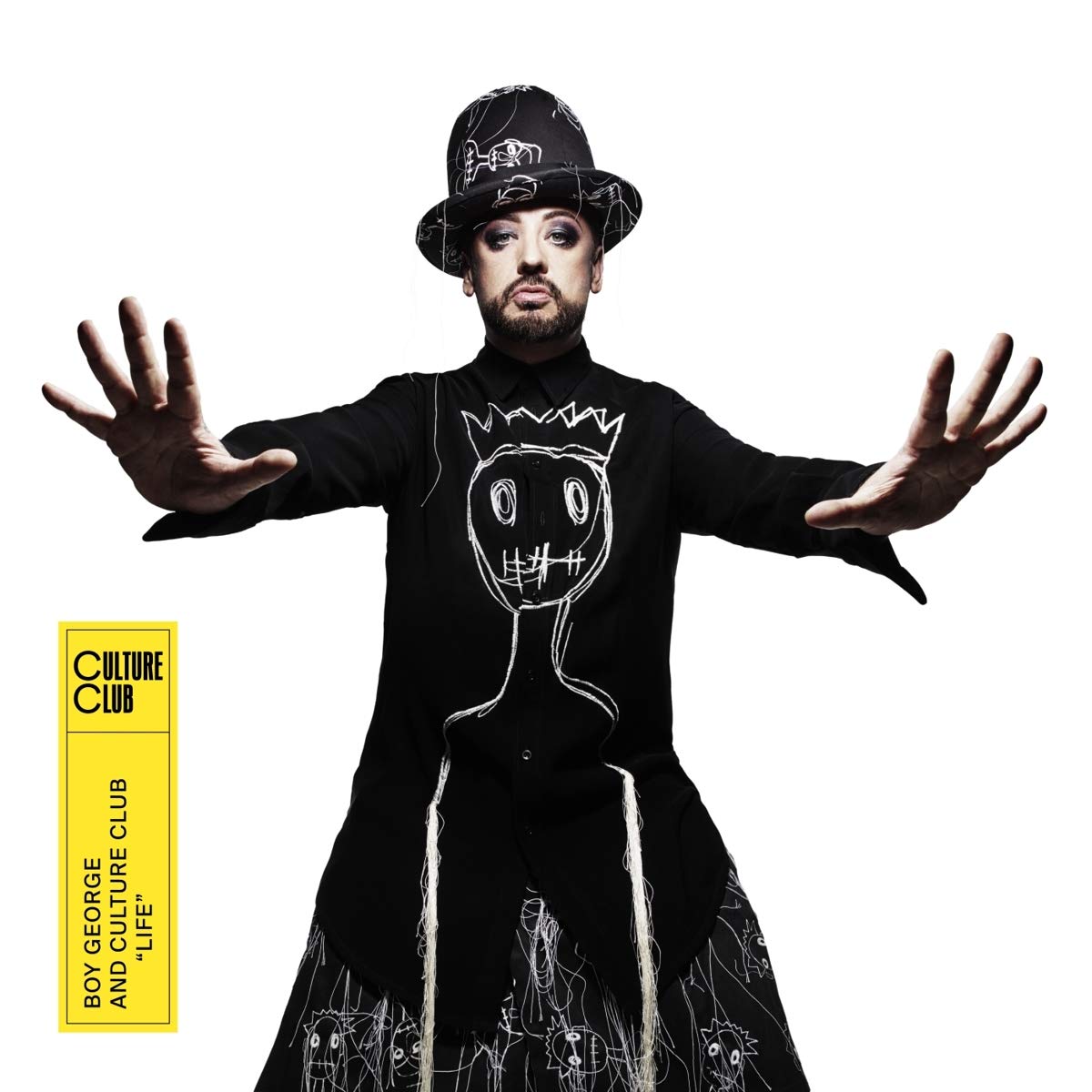 "Boy George in black dress and hat, with outstretched arms, against white background."