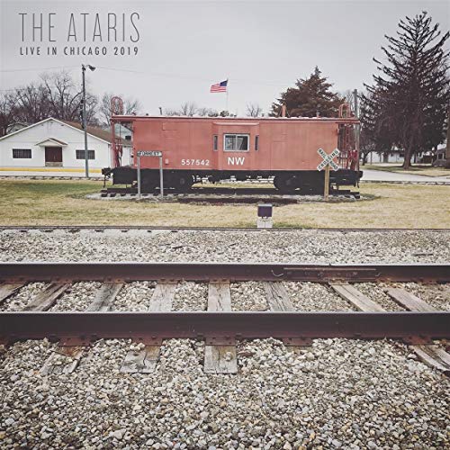 "Red train car on tracks with American flag, trees, and building in background."