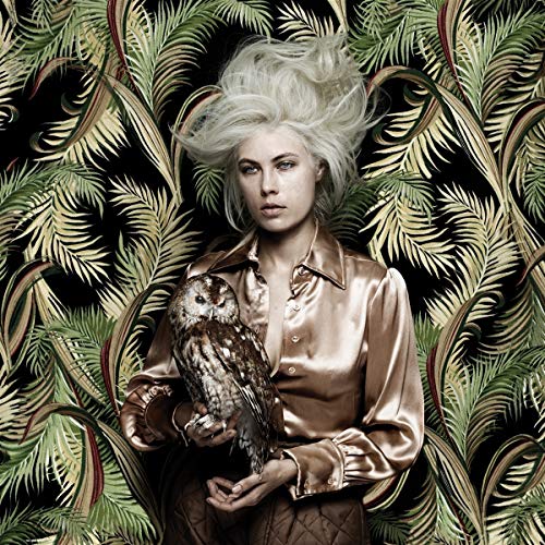 A woman with white hair holds an owl against a leafy background.