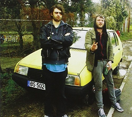 "Two men standing beside a yellow car on a road."