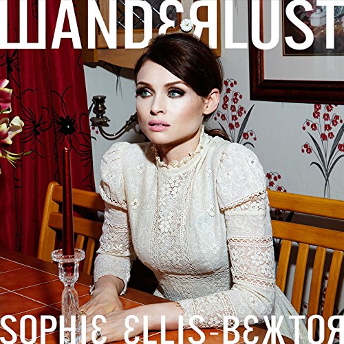 "Sophie Ellis-Bextor in white dress sitting at a wooden table with red curtain."